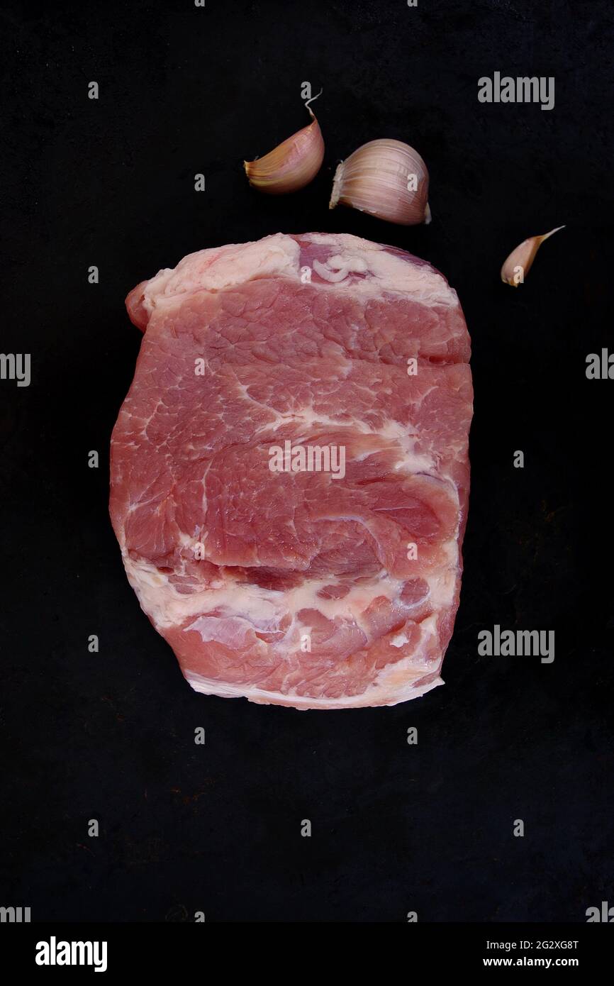 Rohes Stück Schweinefleisch mit rotem frischem Knoblauch auf schwarzem Hintergrund. Ansicht von oben auf ein Stück frisches Schweinefleisch ohne Knochen. Großes Stück rohes Fleisch. Garzeit. Stockfoto