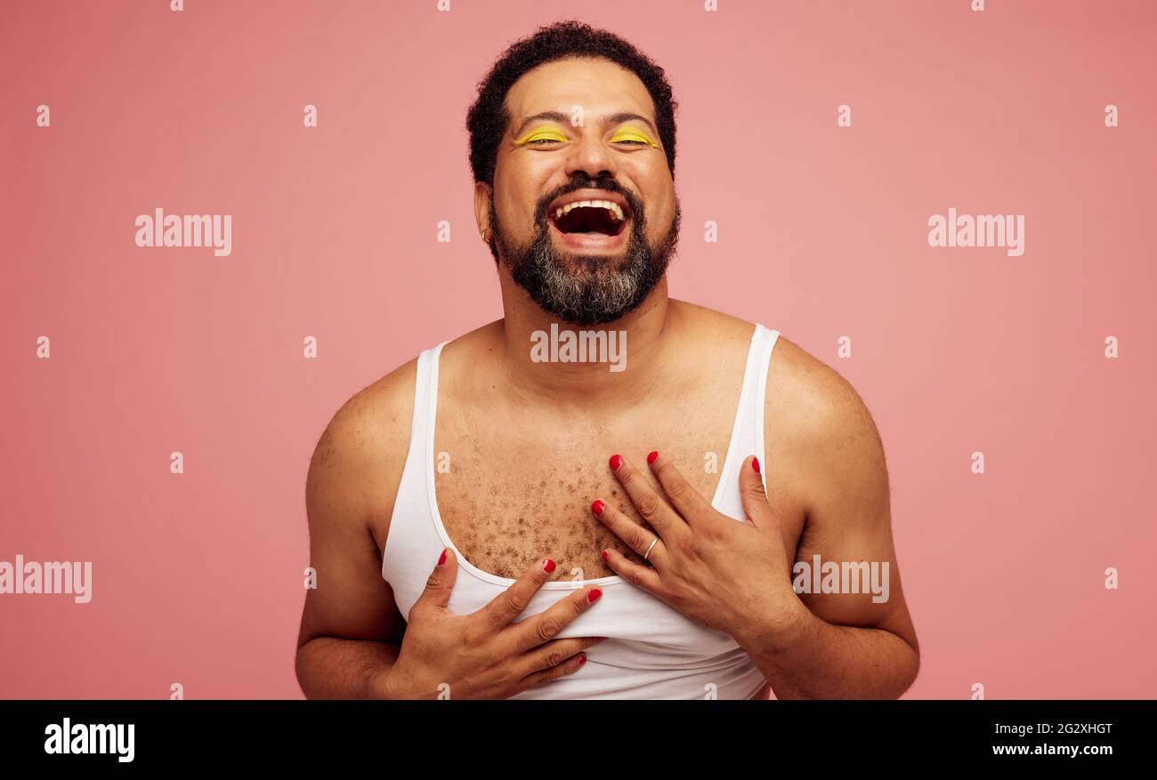 Geschlecht flüssig männlich in einem weiblichen Oberteil und Make-up. Bärtiger Mann mit Lidschatten und Nagellack lachend auf rosa Hintergrund Stockfoto