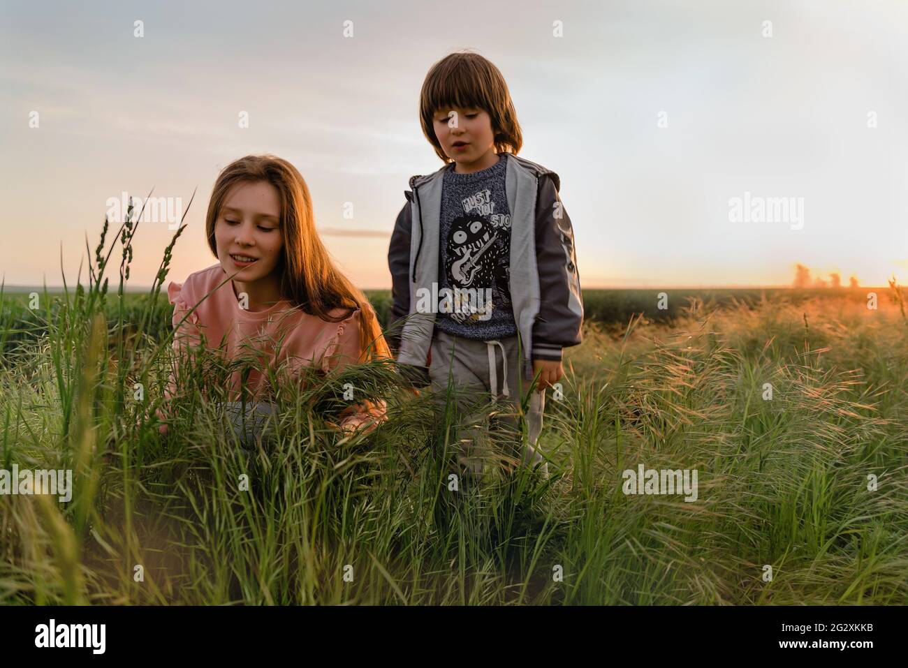 Schönheit junges Mädchen und kleiner Junge, Schwester und Bruder im Freien genießen Natur, Sonnenuntergang im grünen Feld Frühling Stockfoto