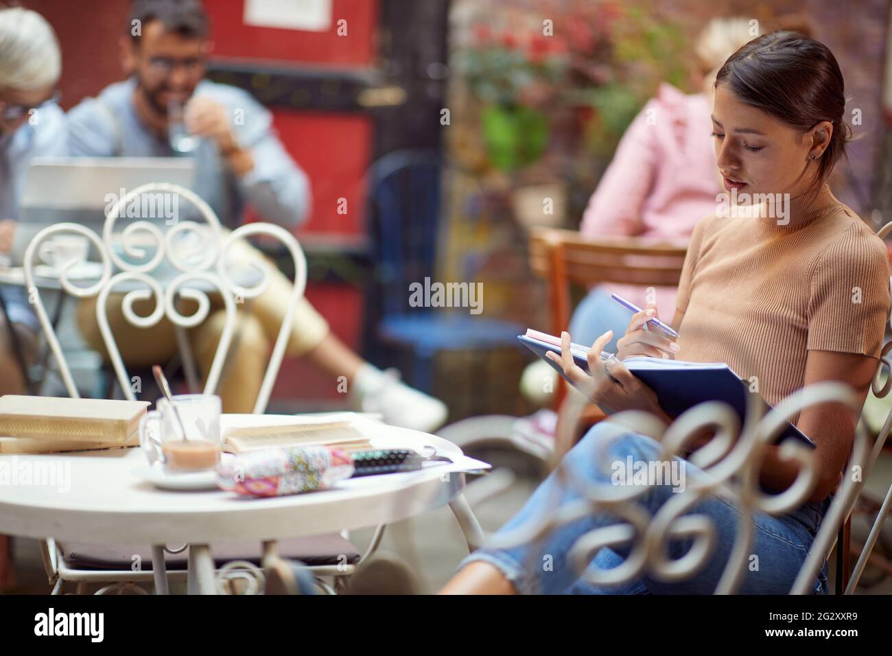 Junge Erwachsene kaukasische Frau, die im Café im Freien sitzt und allein ein Buch liest, mit anderen Menschen im Hintergrund. Stockfoto