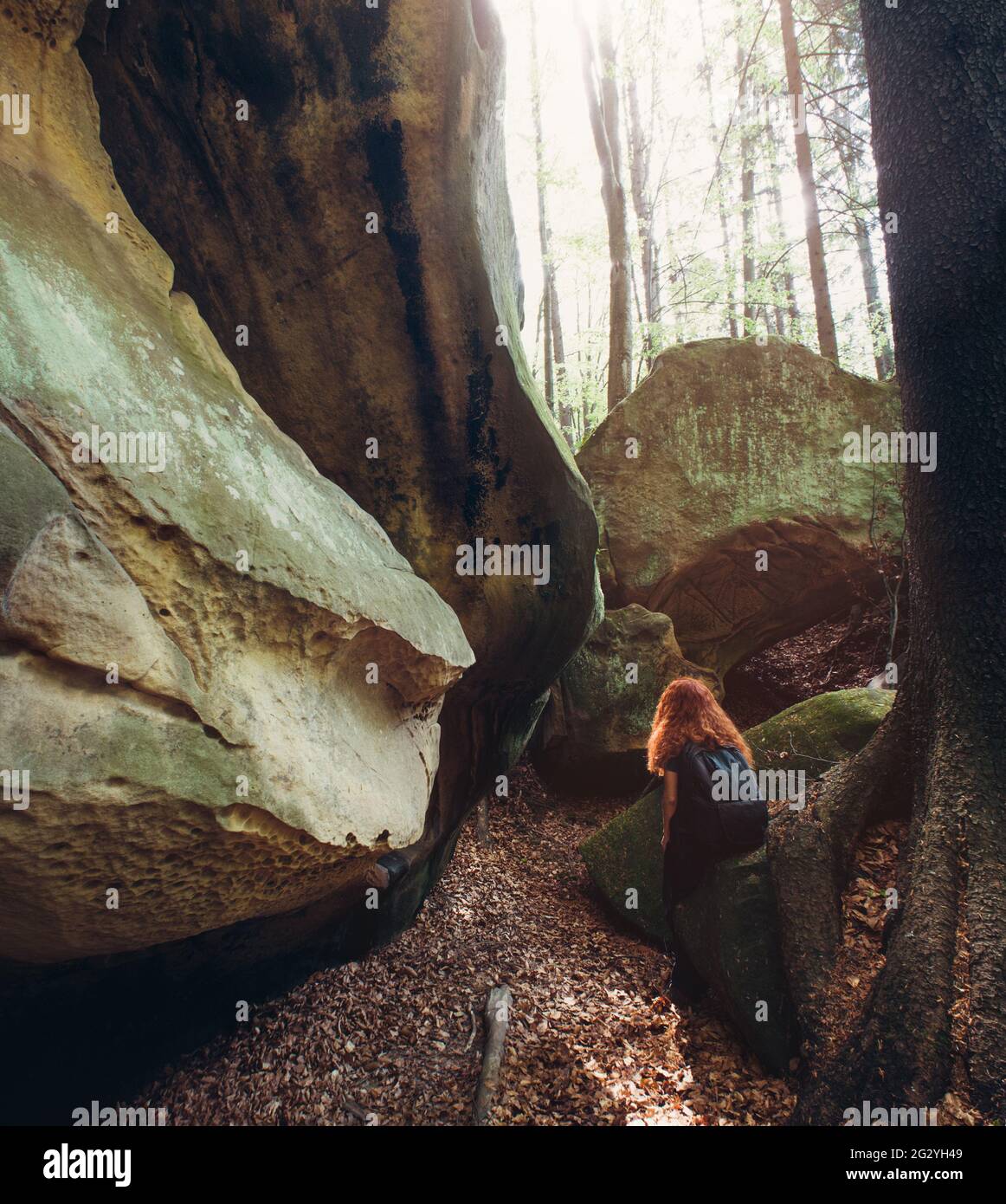 Die rothaarige junge Frau sitzt auf dem riesigen Stein im grünen Frühlingswald und genießt die Aussicht. Konzept des aktiven Lebens. Stockfoto