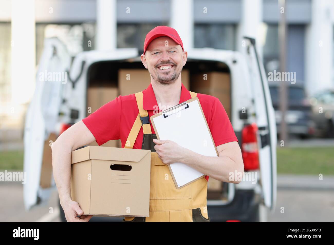 Männlicher Kurier in Uniform hält Klemmbrett und Karton im Hintergrund des LKW mit Boxen Stockfoto