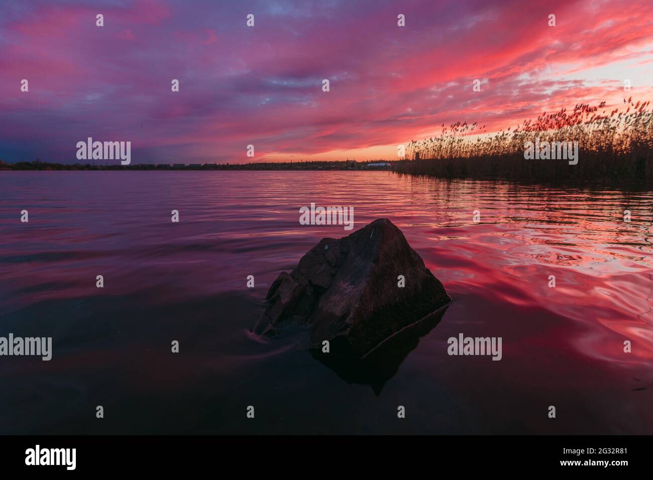 Heller, wolkig kontrastierender, roter Sonnenuntergangshimmel, der sich im Wasser des Sees spiegelte, mit Stein auf der Vorderseite. Das Wasser hinunter Stockfoto