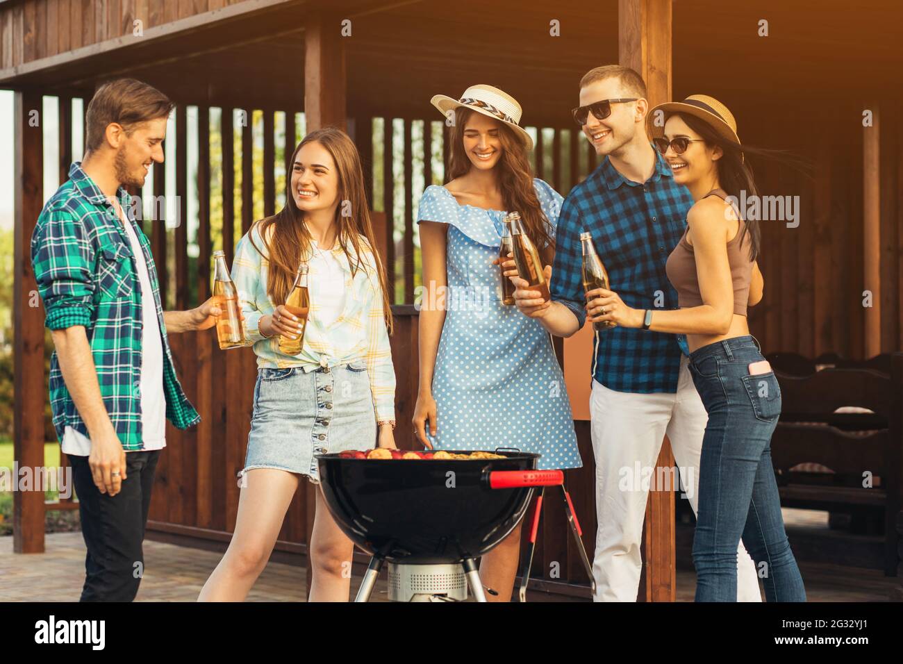 Eine Gruppe von fröhlichen jungen Freunden, die eine Grillparty im Garten machen, Fleisch grillen, trinken und sich an einem sonnigen Sommertag im Freien entspannen Stockfoto