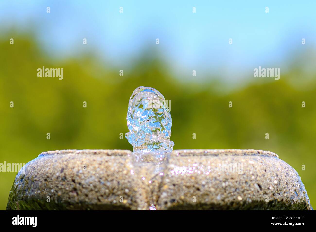 Eine Nahaufnahme eines Steintrinkbrunnens im Freien. Sehr geringe Schärfentiefe, nur Wasser im Fokus, aber Bäume dahinter und blauer Himmel darüber. Platz für Text. Stockfoto