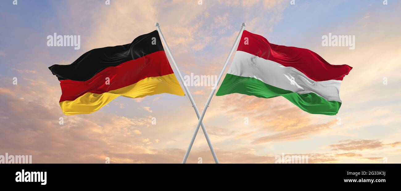 Flaggen von Deutschland und Ungarn winken im Wind auf Fahnenmasten gegen den Himmel mit Wolken am sonnigen Tag. Symbolisieren Beziehung, Dialog zwischen zwei Gräfin Stockfoto