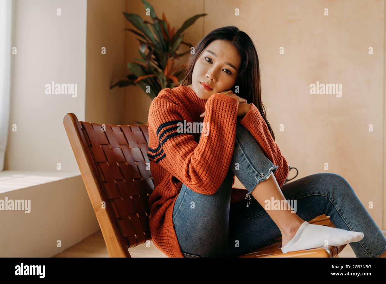 Einsame junge asiatische Frau im Pullover, die sich am Knie lehnt und die Kamera anschaut, während sie sich auf einem Stuhl im gemütlichen Zimmer zuhause ausruht Stockfoto