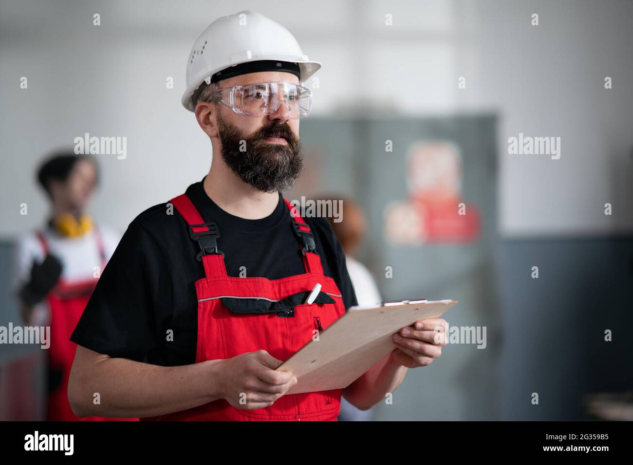 Portrait eines Arbeiters mit Helm und Schutzbrille im Innenbereich in der Fabrik mit Klemmbrett. Stockfoto