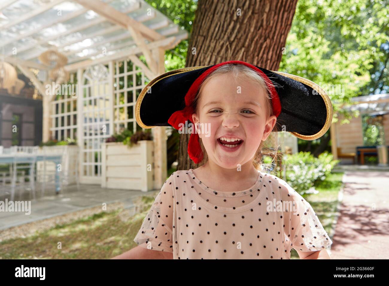 Entzückendes Mädchen im Piratenhut mit Blick auf die Kamera und lachen, während sie im Sommer im Innenhof Spaß haben Stockfoto