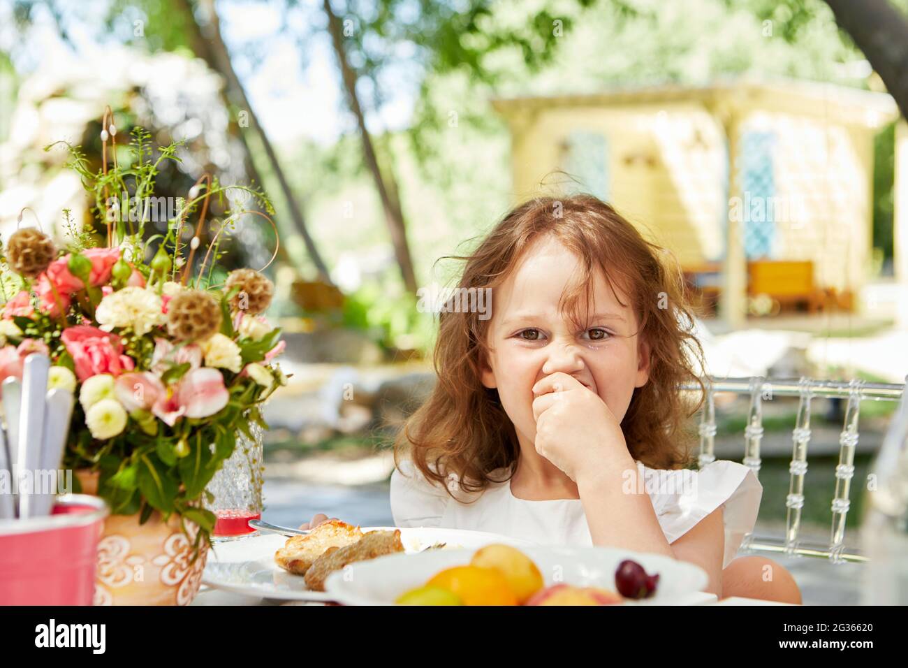 Nettes kleines Kind, das leckeres Essen isst, während es am Tisch im Hinterhof sitzt und die Kamera anschaut Stockfoto