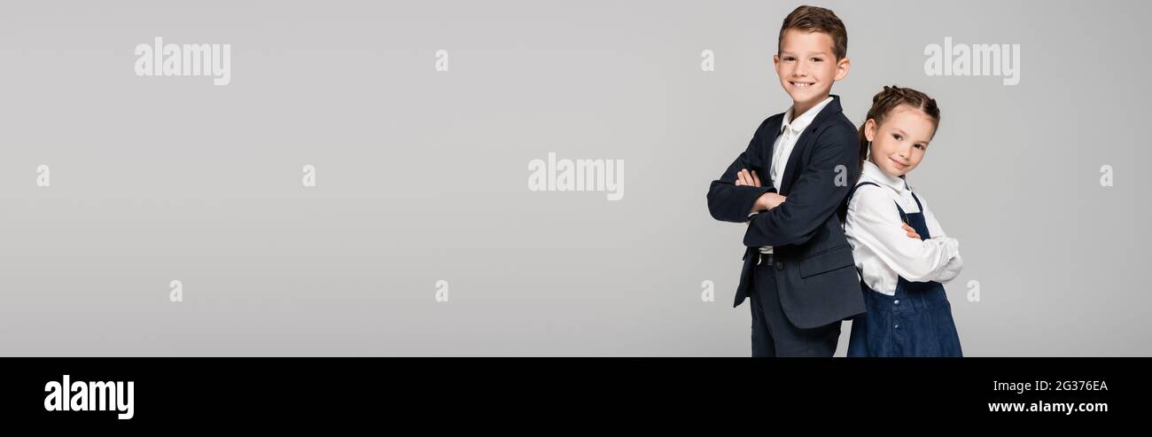 Glückliche Schüler in Uniform posieren mit gekreuzten Armen isoliert auf grauem Banner Stockfoto