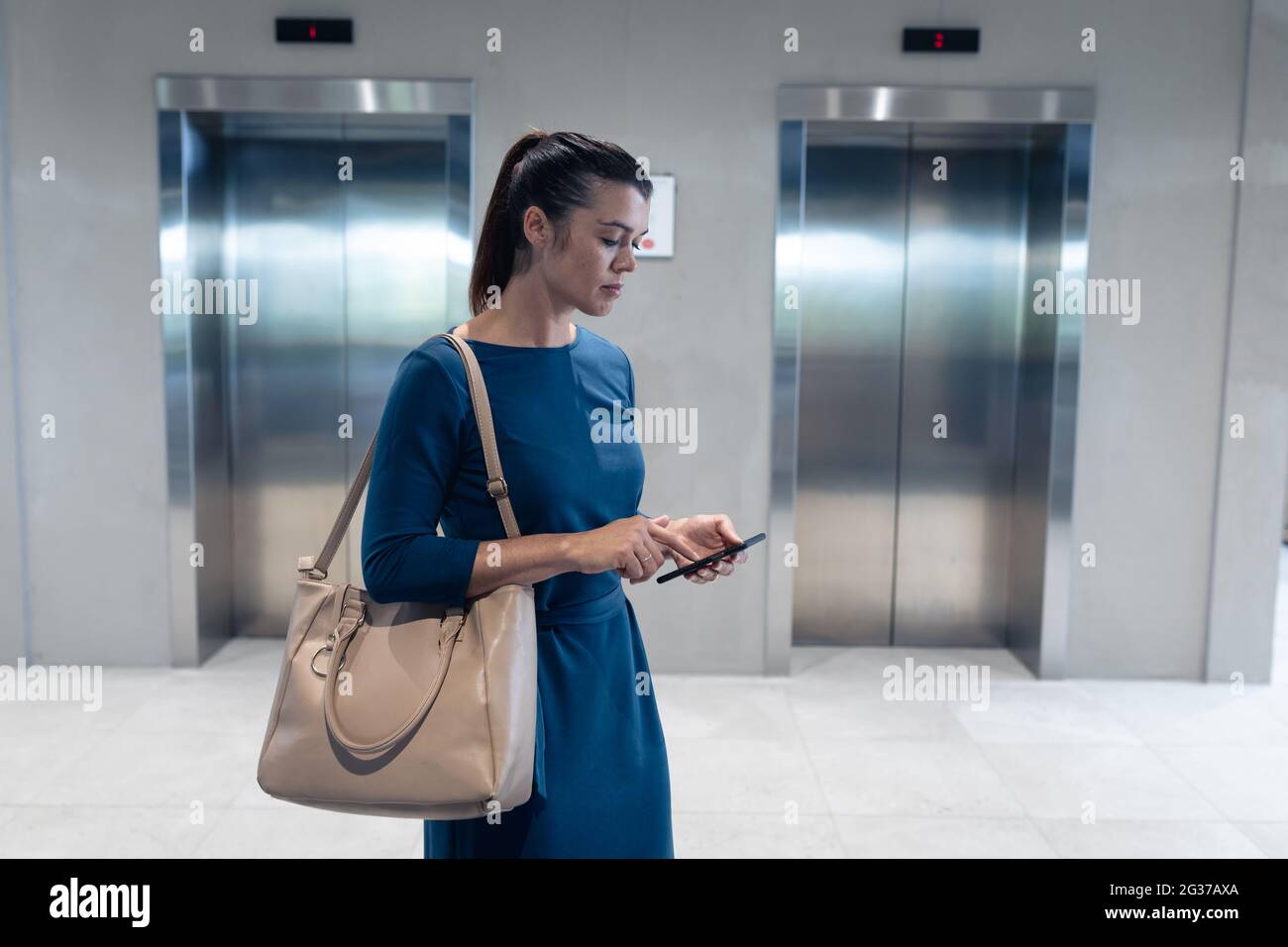 Kaukasische Geschäftsfrau mit Smartphone, während sie auf den Aufzug im modernen Büro wartet Stockfoto
