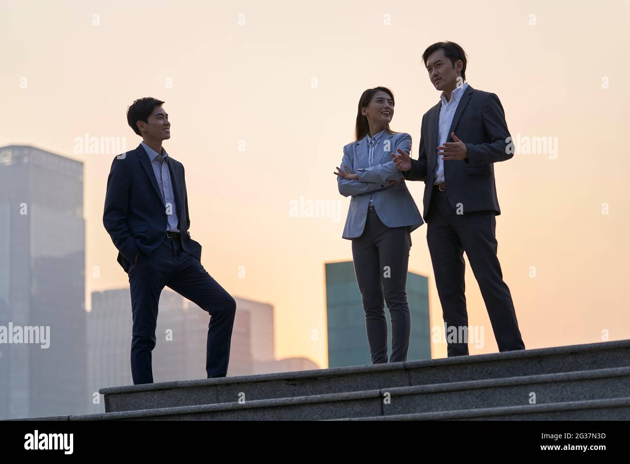 Team von asiatischen Unternehmensleitern, die auf einer Plattform stehen und sich mit Gesprächen unterhalten, die in der Abenddämmerung mit Stadthintergrund unterhalten Stockfoto