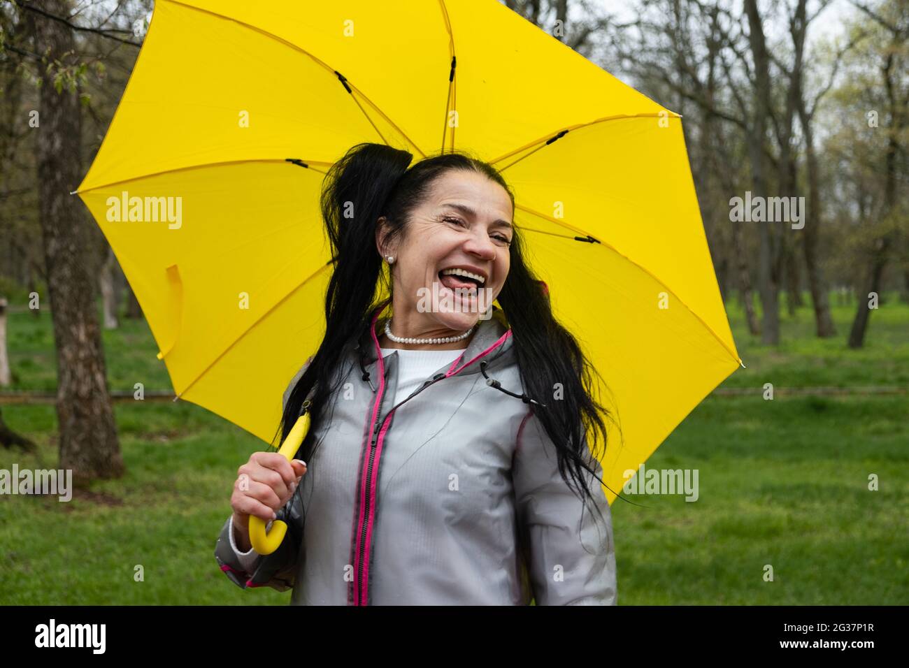 Glückliche ältere fröhliche ältere, ältere, pensionierte Frau mit gelbem Regenschirm, die das Leben an regnerischen Tagen im Park genießt. Genießen Sie jeden Moment, genießen Sie das Leben Stockfoto