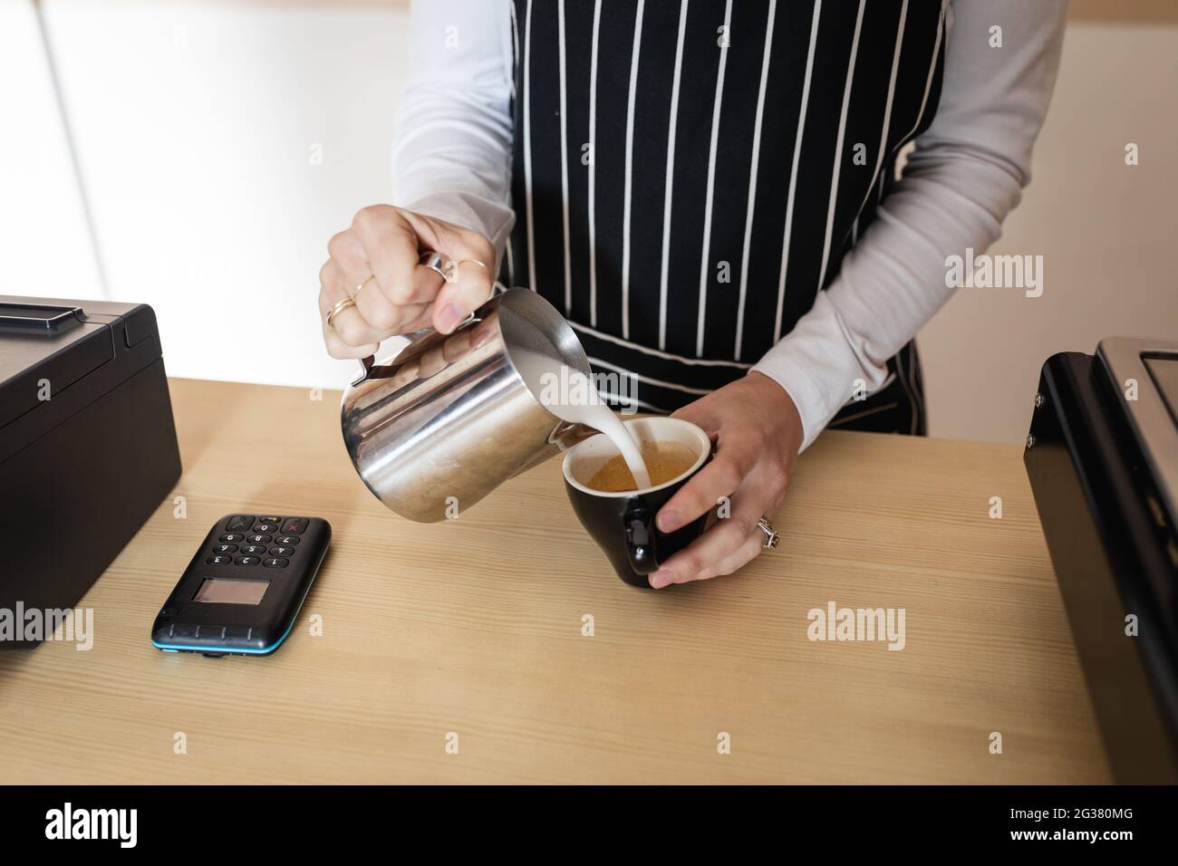 Mittelteil der kaukasischen Geschäftsbesitzerin, die Schürze trägt und Milchschaum in den Kaffee gießt Stockfoto