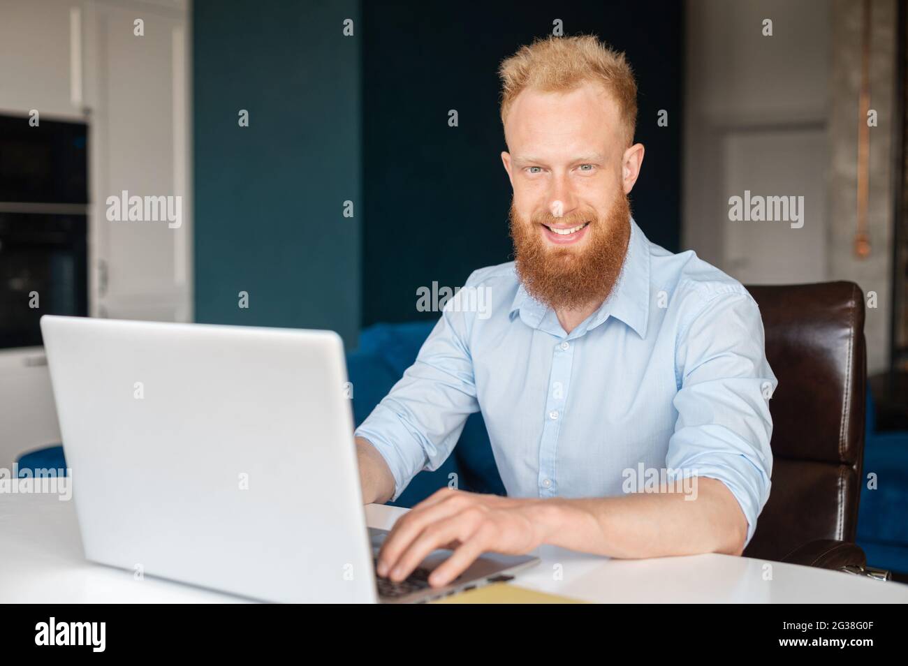 Der ehrgeizige, rothaarige, bärtige Unternehmer blickt mit einem freundlichen Lächeln auf die Kamera, ein junger bärtiger Mann im Hemd sitzt am Schreibtisch und benutzt einen Laptop in der Halle, einen männlichen Büroangestellten Stockfoto