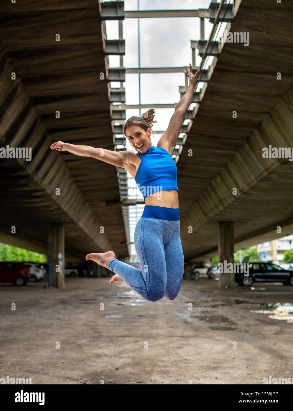 Sportliche Frau lächelt glücklich beim Training im Freien auf dem Parkplatz. Sportliche Mädchen springen mit den Armen nach oben und die Beine gebeugt. Stockfoto