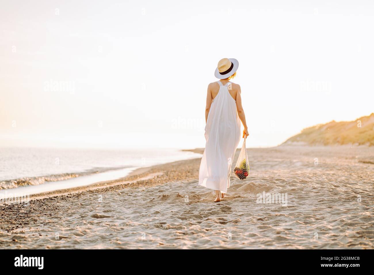 Wiederverwendbares, recycelbares Lifestyle-Konzept. Frau in Naturkleidung geht an einem Strand entlang und hält wiederverwendbare Mesh-Einkaufstasche mit Gemüse Stockfoto