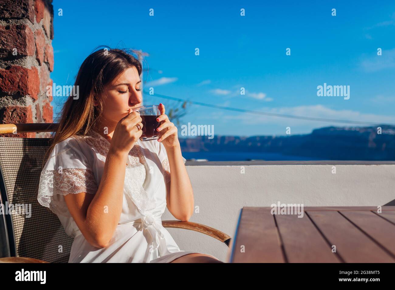 Frau, die sich auf der Hotelterrasse entspannt, trinkt morgens Kaffee und genießt die Berg- und Meereslandschaft. Sommerurlaub Stockfoto