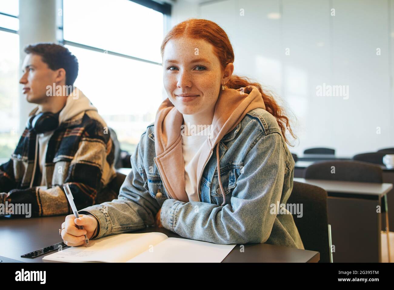 Schöne Schülerin lächelt zur Kamera, während sie Notizen im Klassenzimmer schreibt. Junge Frau, die ihre Klasse besucht. Stockfoto