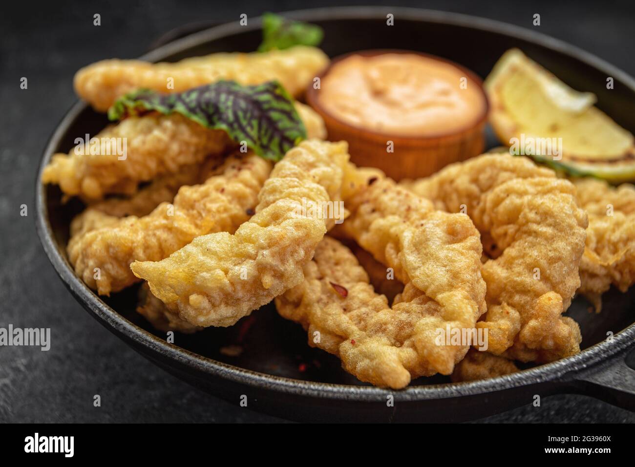 Goldene Ringe von Tintenfisch-Pommes in einer runden Bratpfanne mit Sauce, Kräutern und Zitrone auf dunklem Hintergrund ohne Verzierungen Stockfoto