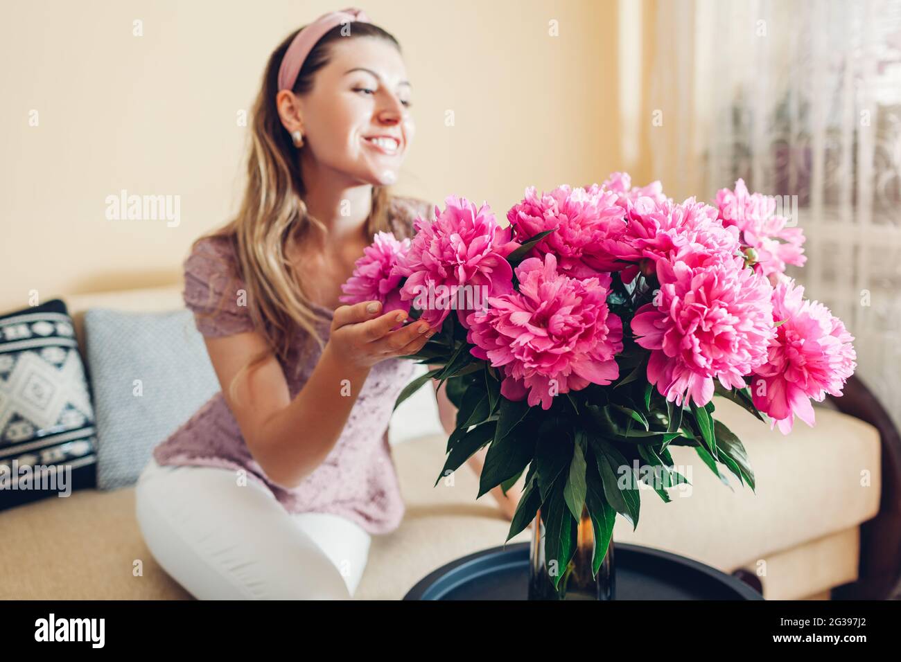 Frau bewundert zu Hause einen Strauß rosa Pfingstrosen in der Vase. Mädchen riecht blüht. Innenausstattung und gemütliche Wohnzimmereinrichtung Stockfoto
