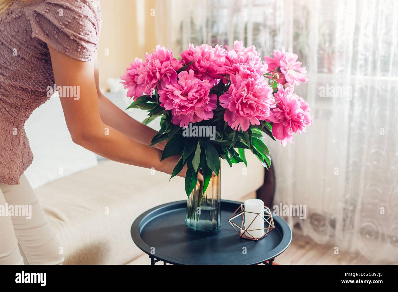 Frau stellt Vase mit Blumenstrauß aus rosa Pfingstrosen auf den Tisch. Hausfrau kümmert sich um Gemütlichkeit zu Hause. Einrichtung und Einrichtung im Frühling Stockfoto
