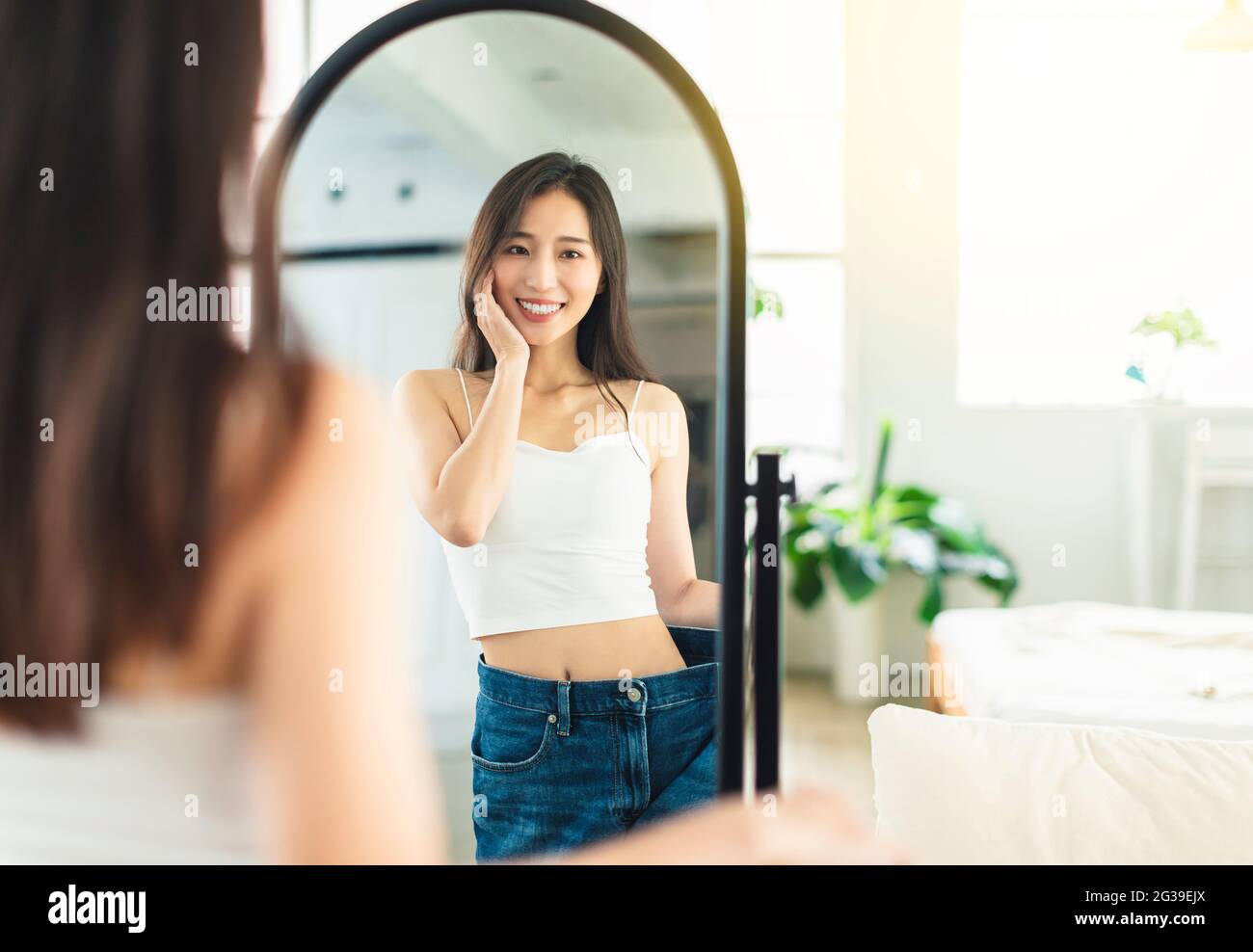 Die junge Frau, die vor dem Abnehmen große Jeans trägt und die Figur vor dem Spiegel anschaut, ist sehr glücklich, dass sie erfolgreich abgenommen hat Stockfoto