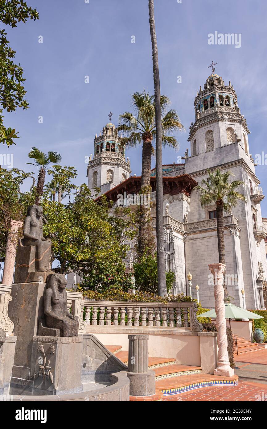 San Simeon, CA, USA - 12. Februar 2014: Hearst Castle. Türme und formelle Eingangsfassade hinter grünem Laub und schwarzen ägyptischen Statuen unter blauen s Stockfoto