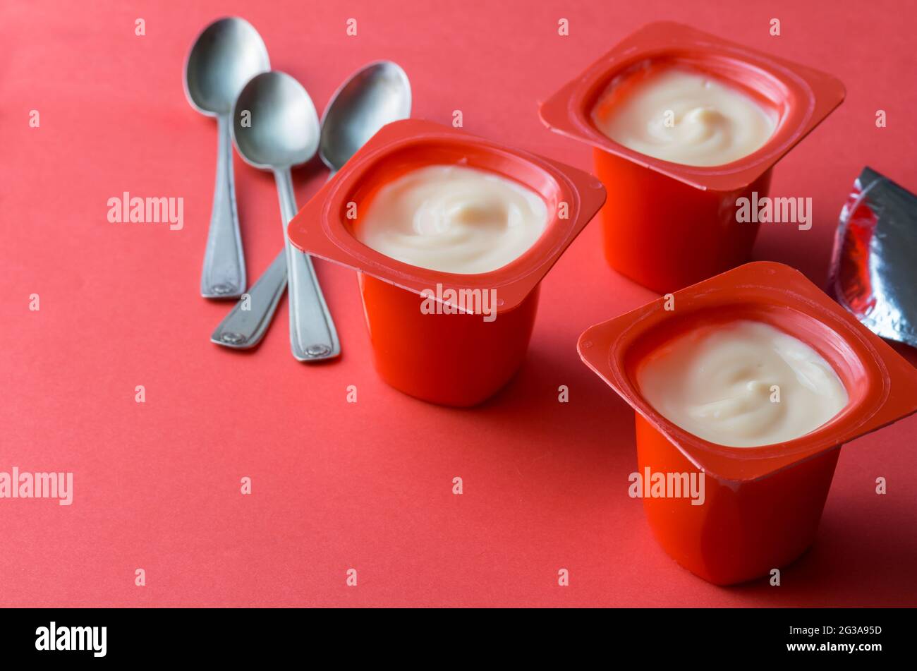 Drei rote Joghurt-Plastiktöpfe mit Löffeln und Folie auf rotem Hintergrund - cremiger Fruchtjoghurt in Plastikbechern mit Kopierfläche Stockfoto