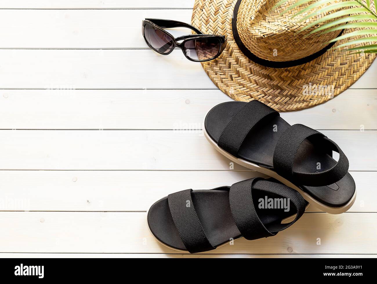 Schwarze Sandalen mit offenem Zehenbereich aus recycelten Kunststofffasern, Strohhut und Sonnenbrille auf der Holzpromenade. Reisekonzept für den Sommer. Stockfoto