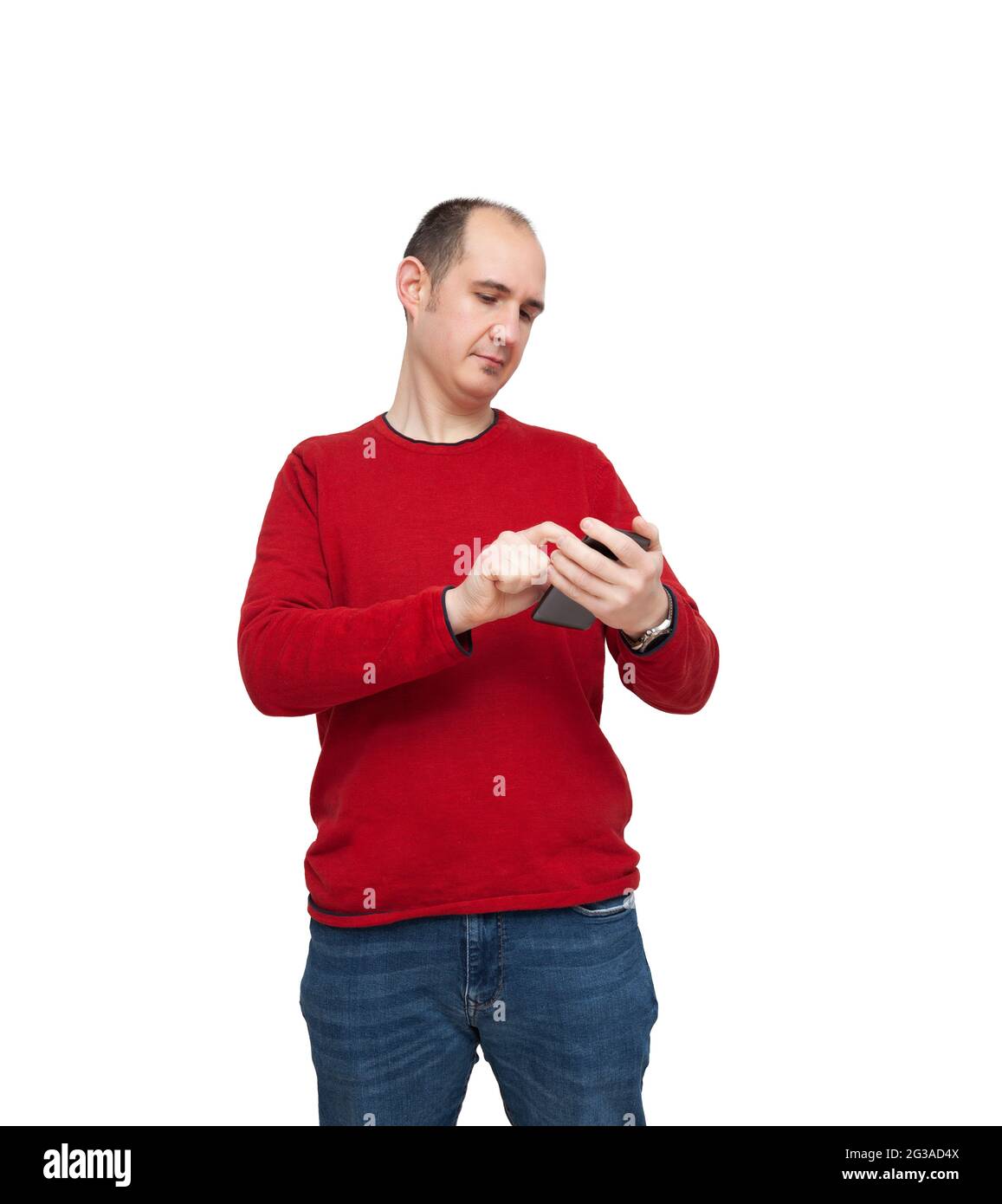 Ein kahler junger Mann tippt mit seinem Mobiltelefon eine Nachricht ein. Die Person ist mit einem roten Pullover, Jeans und einer Uhr bekleidet. Der Hintergrund ist weiß. Stockfoto