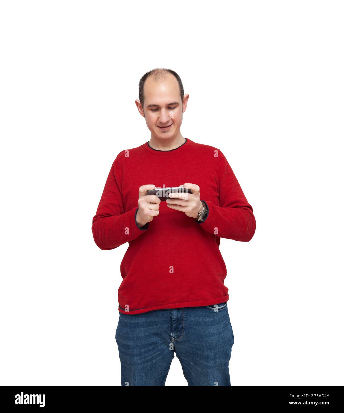 Ein kahler junger Mann spielt mit seinem Mobiltelefon ein Videospiel. Die Person ist mit einem roten Pullover, Jeans und einer Uhr bekleidet. Der Hintergrund ist Whi Stockfoto