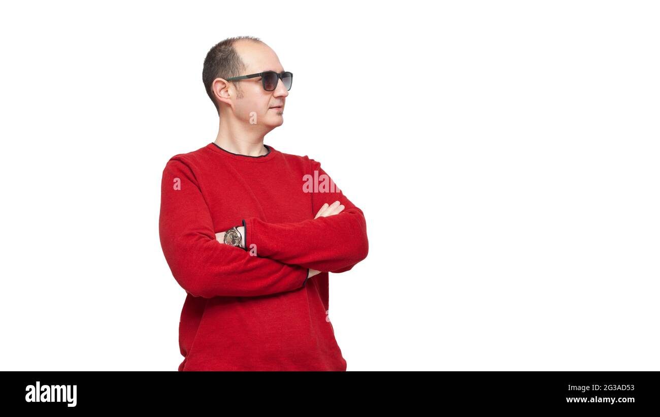 Ein kahler junger Mann, der in einem roten Pullover und einer Sonnenbrille gekleidet ist, steht mit gekreuzten Armen auf der rechten Seite des Bildes. Der Hintergrund ist weiß. Stockfoto