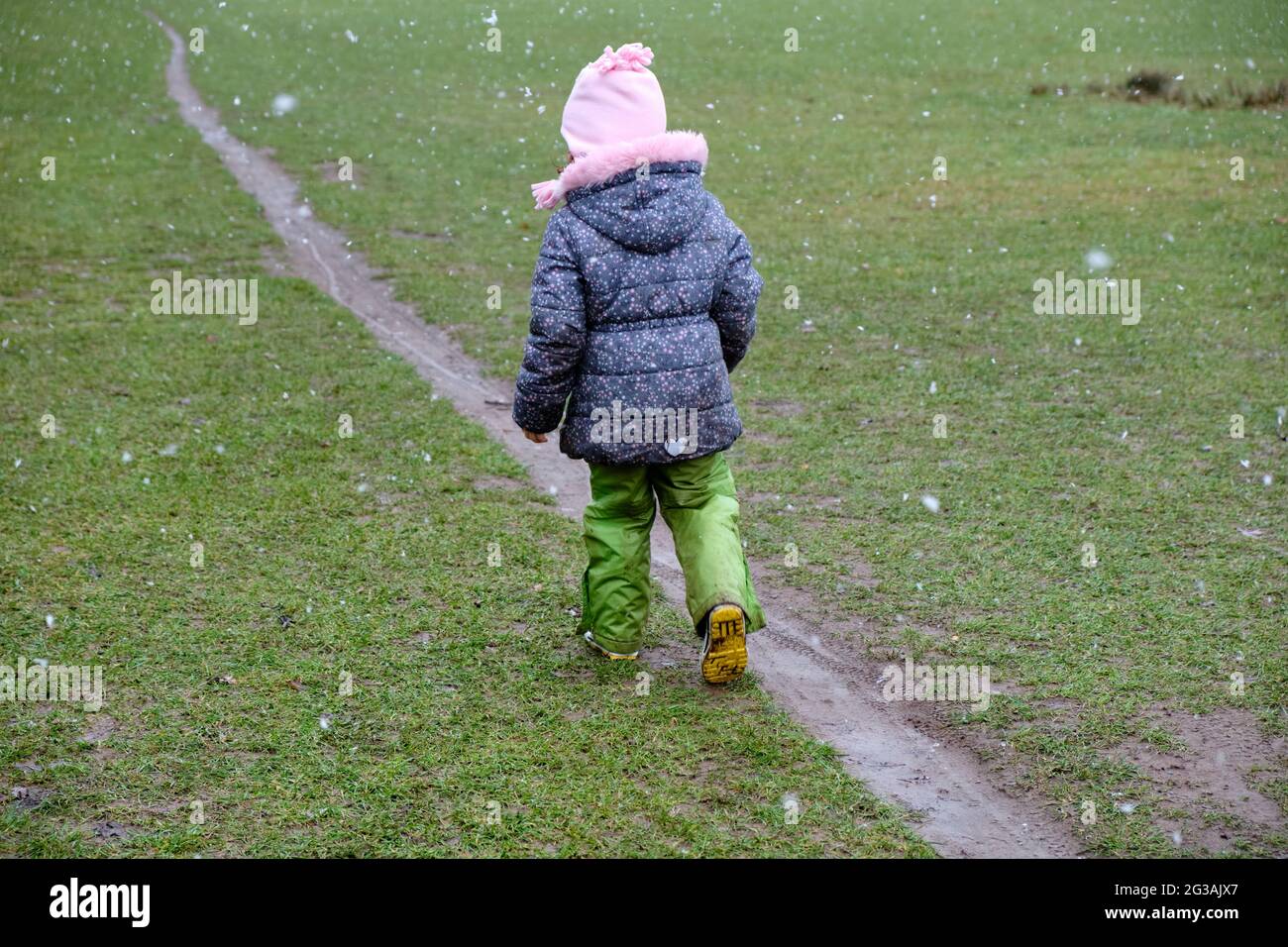 Rückansicht eines 5-jährigen Mädchens in warmer Kleidung, das im Winter während des Schneefalls auf einer Wiese auf einem schmalen, schlammigen Pfad läuft. Gesehen in Deutschland in Janua Stockfoto