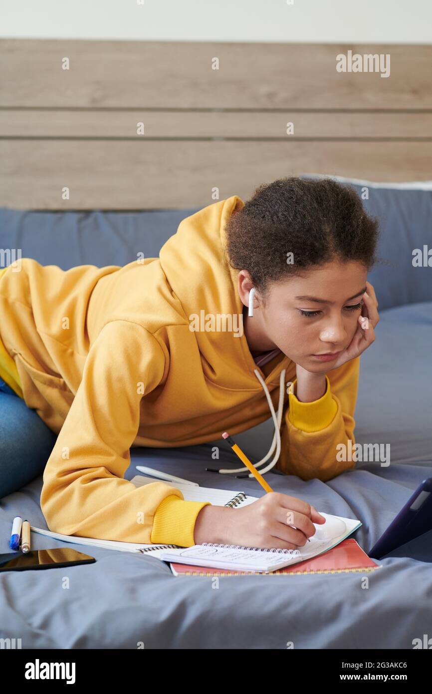 Unzufrieden gemischtes High-School-Mädchen in drahtlosen Kopfhörern, die auf dem Bett liegen und Notizen machen, während sie die Schulaufgabe lösen Stockfoto