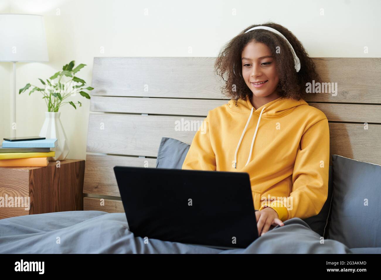 Content Mixed Race Girl in kabellosen Kopfhörern, die auf dem Bett sitzen und auf dem Laptop tippen, während sie im Internet nach Informationen suchen Stockfoto