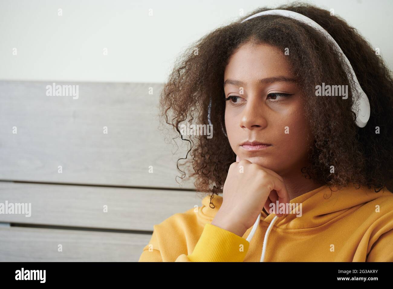 Trauriges, nachdenkliches Teenager-Mixed-Race-Girl mit lockigen Haaren, das Musik in weißen Kopfhörern hört und den Kopf an der Hand lehnt Stockfoto