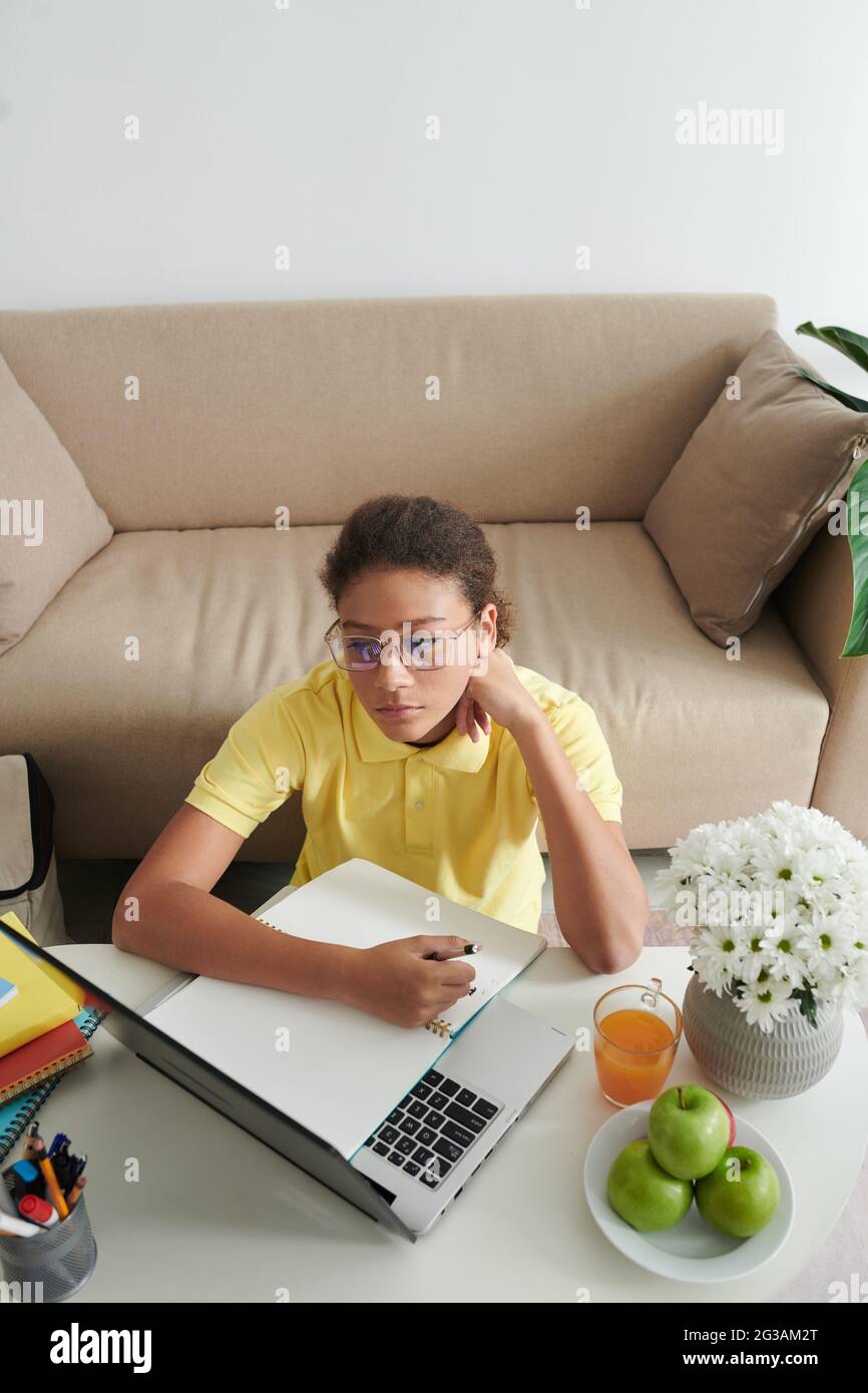 Serious clever Mixed-Race Teenager Mädchen in Brillen sitzen am Couchtisch im Wohnzimmer und mit Laptop, während Online-Unterricht Stockfoto