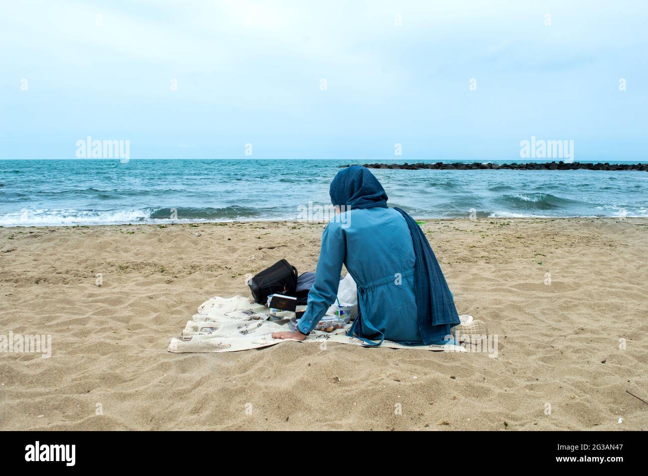 Hijab-Mädchen in blauem Turban macht ein Picknick am Strand. Frau in Hijab sitzt am Strand. Picknick am Strand. Picknick und Freizeit Sehenswürdigkeiten. Stockfoto