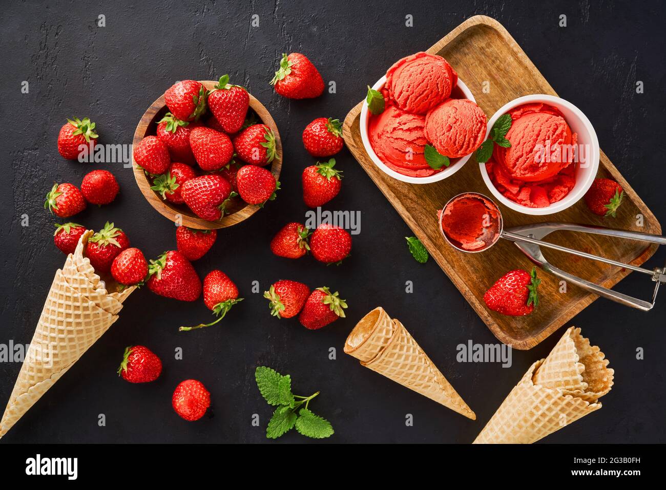 Erdbeerhaufen mit Eis in Eiscreme-Pappbechern und -Zapfen. Erdbeersorbet auf schwarzem Hintergrund. Stockfoto