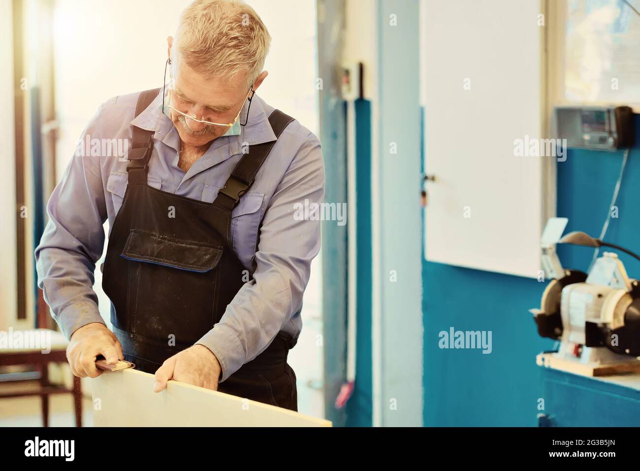 Porträt eines älteren Zimmermanns oder Zimmermanns in Overalls, der in einer Tischlerwerkstatt mit Holzbrettern arbeitet. Sägen von Holz. Stockfoto