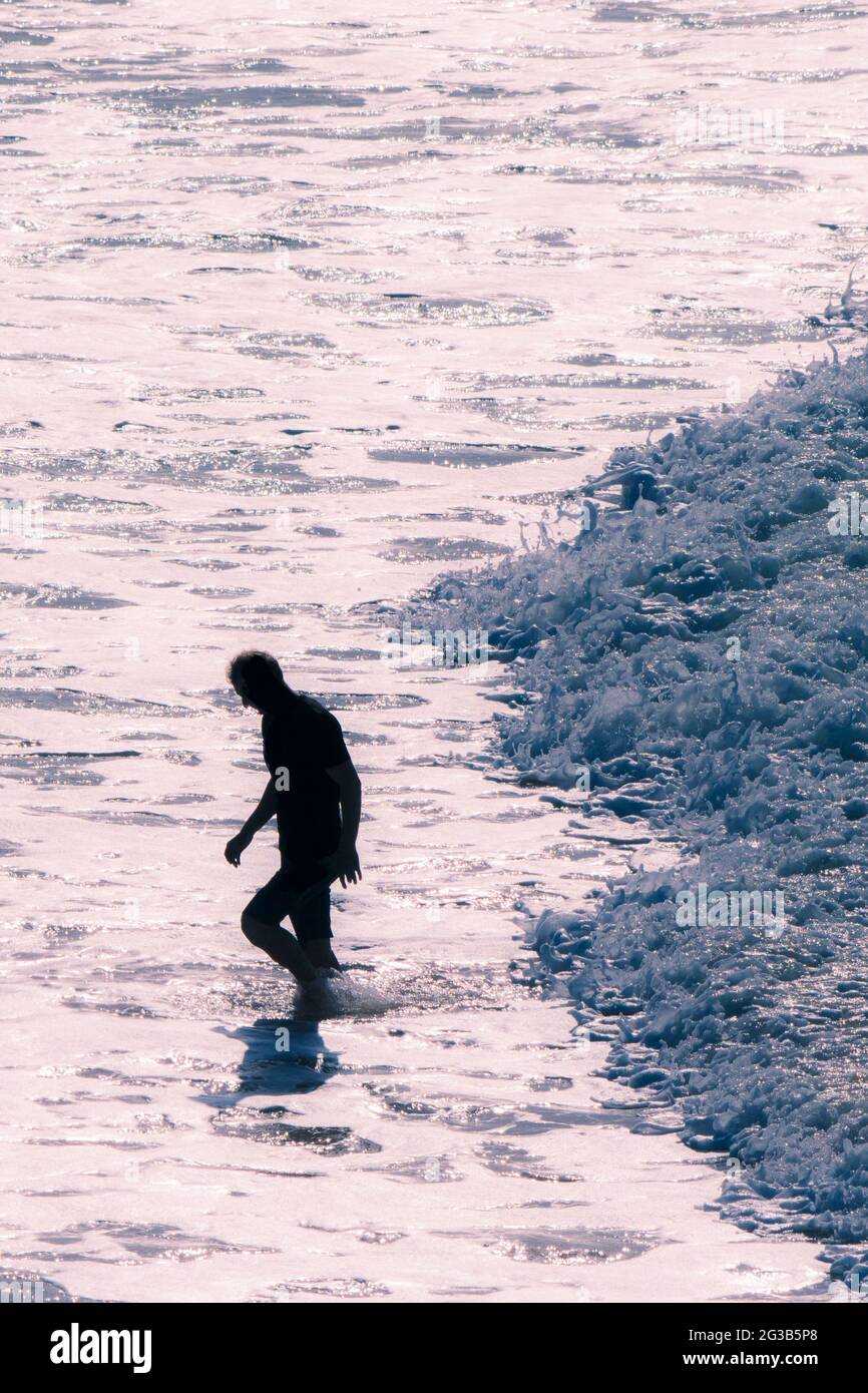 Ein Mann, der durch das Abendlicht aus dem Meer hinausläuft, gefolgt von einer kleinen Welle, die sanft am Fistral Beach in Newquay in Cornwall fließt. Stockfoto