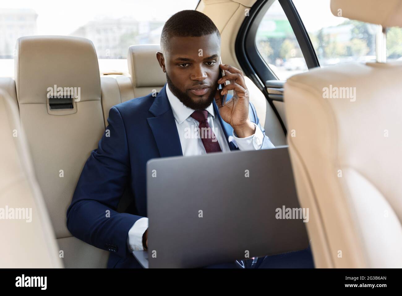Wohlhabender afroamerikanischer Geschäftsmann in stilvollem Anzug, der im Auto arbeitet, Laptop benutzt und auf dem Smartphone spricht, online arbeitet, Speicherplatz kopiert. Konzentriert Stockfoto