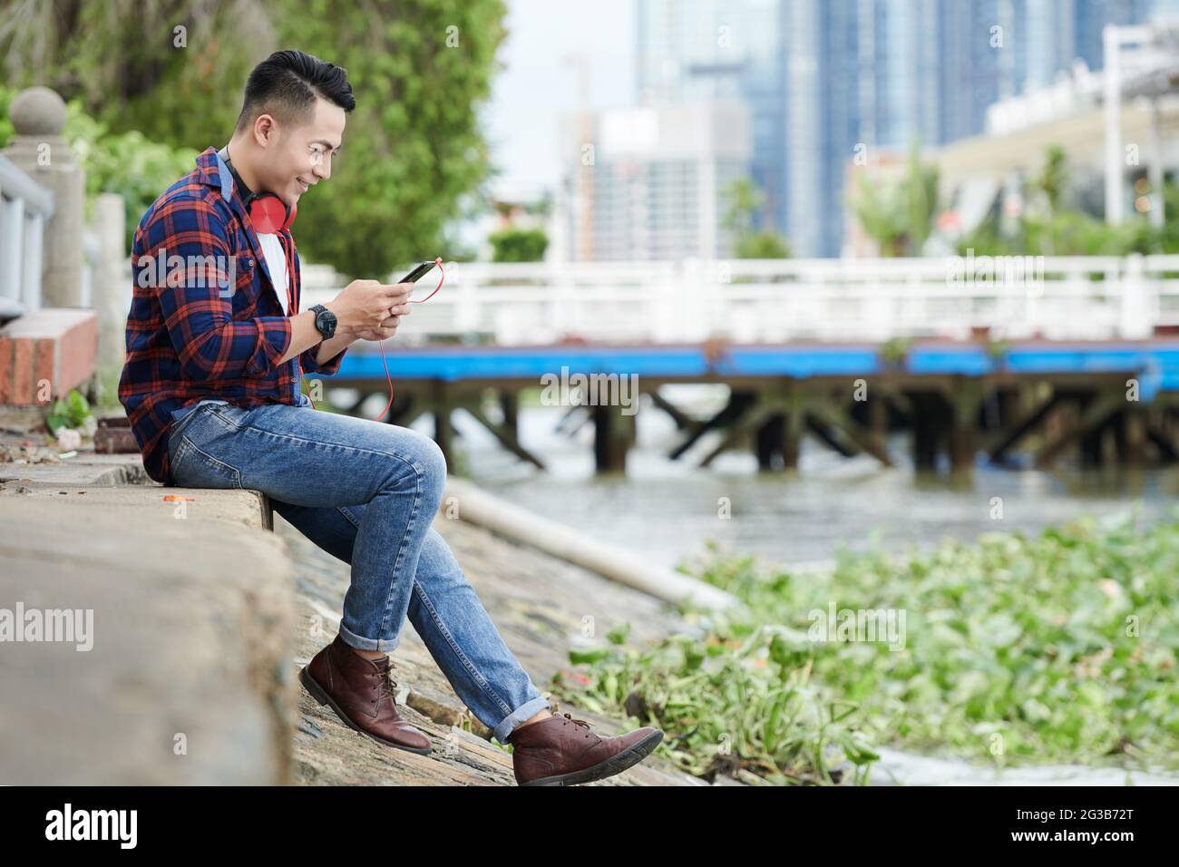 Lächelnder junger vietnamesischer Mann im lässigen Outfit sitzt auf dem Boden und schreibt eine Nachricht auf dem Smartphone, während er online chattet Stockfoto