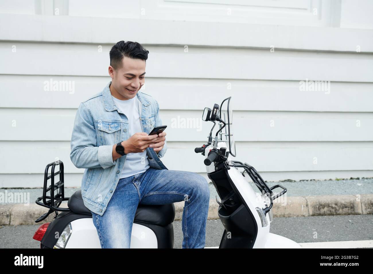 Lächelnder junger vietnamesischer Moped-Fahrer im Denim-Outfit sitzt auf einem modernen Fahrrad und nutzt das Smartphone, während er online kommuniziert Stockfoto