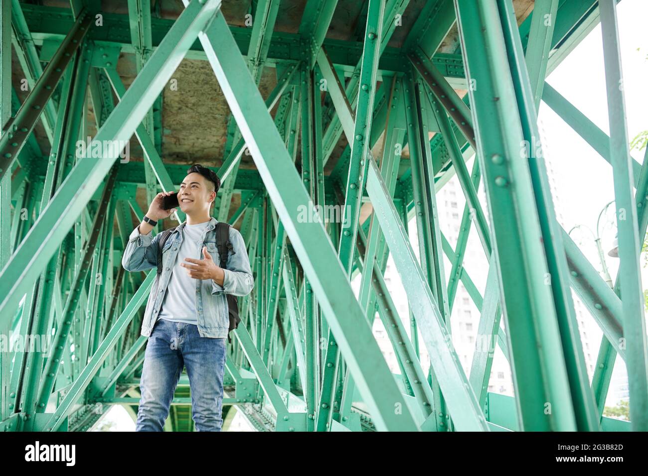 Positiver junger vietnamesischer Tourist im Denim-Outfit, der unter einer Brücke mit grünen Metallträgern steht und die Hand während des Telefonats gestikelt Stockfoto