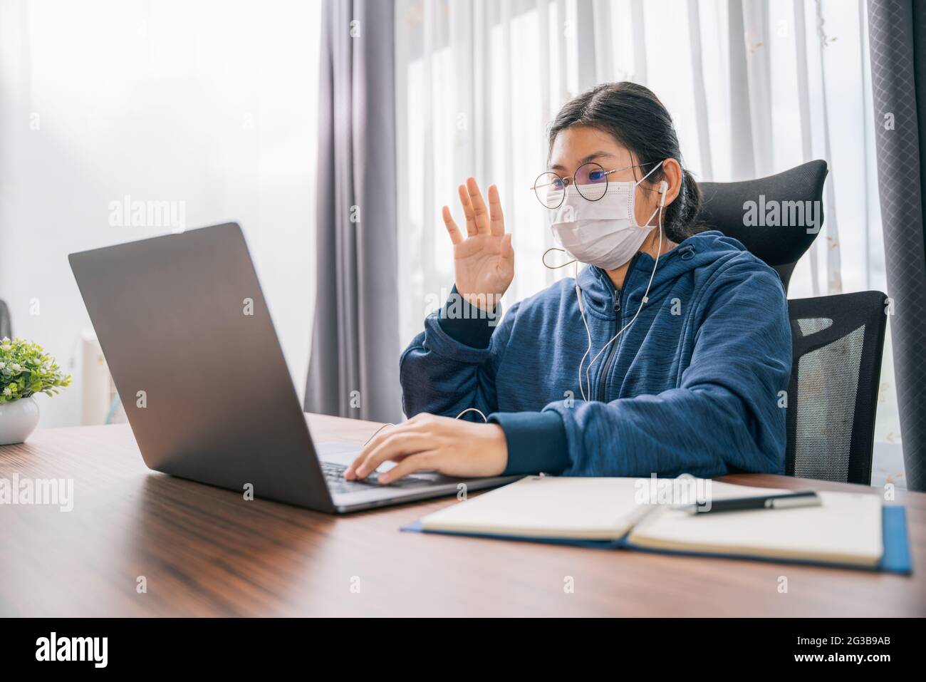 Asiatische junge Mädchen tragen eine Maskengeste der Begrüßung Student Lernen online mit Kopfhörern und Laptop zu Hause Stockfoto