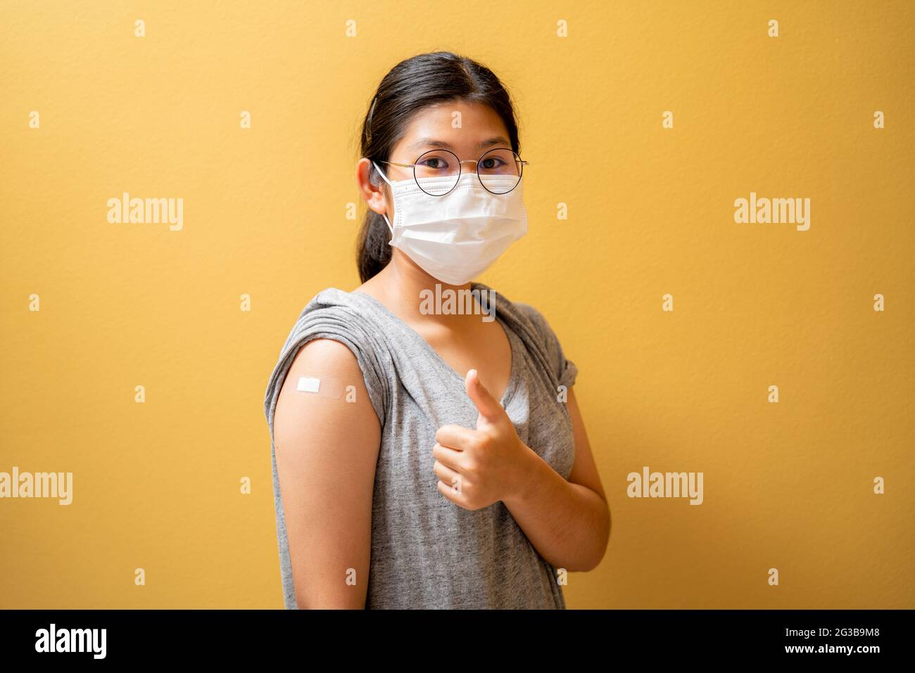 Geimpftes asiatisches Teenager-Mädchen in Gesichtsmaske, das Gipsverband auf ihrem Arm zeigt, nachdem es eine covid-19-Impfstoffinjektion auf gelbem Studiohintergrund erhalten hatte. Coron Stockfoto