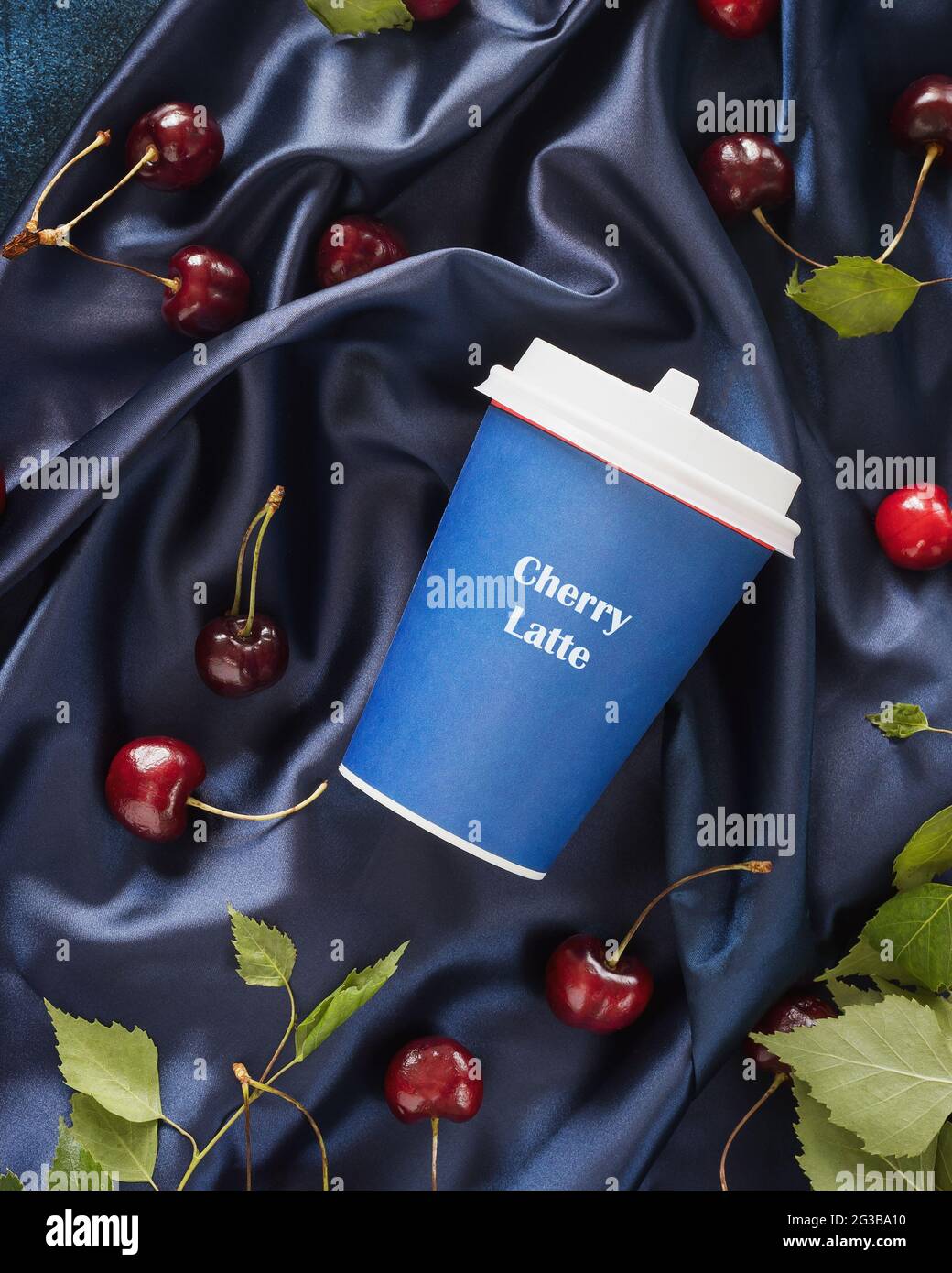 Cherry Latte, eine neue Art von Kaffee. Lebensmittelkomposition mit einer Tasse Kaffee, Kirschen und Blättern auf einem blauen, satinierten, gefalteten Hintergrund. Kaffee zum trinken Stockfoto